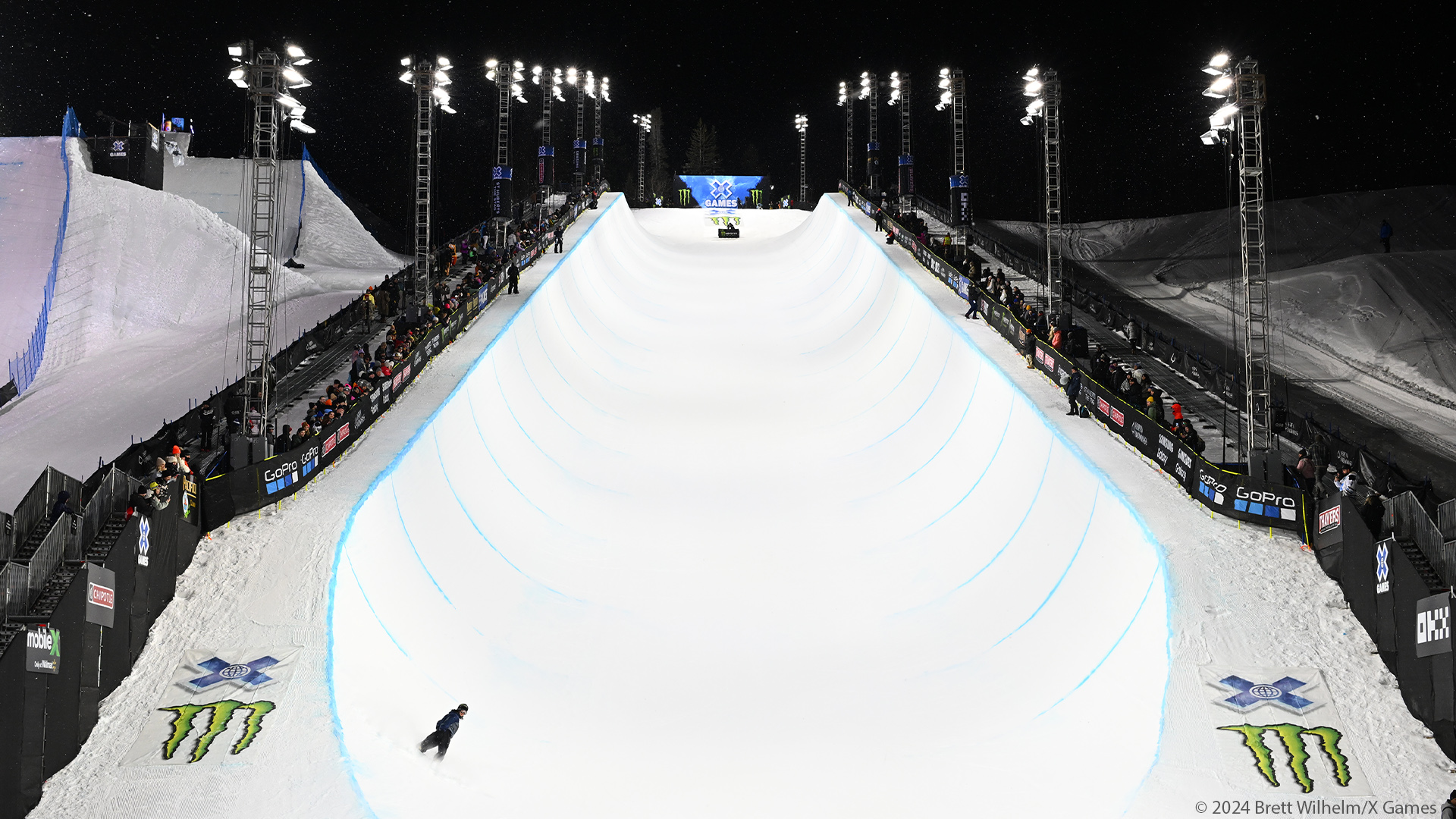 Snowboarder finishes night run on X Games Superpipe at 2024 winter games in Aspen, A row of Musco LED temporary lighting lines the Superpipe on either side.