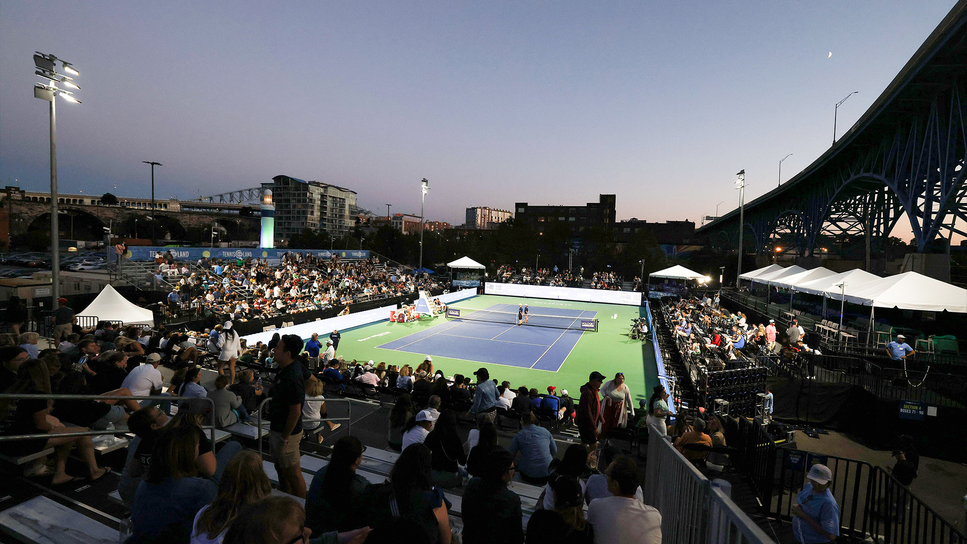 Blue and green tennis court at Tennis in the Land surrounded by people in the stands and lit at sunset with Musco temporary lighting system with LED fixtures.
