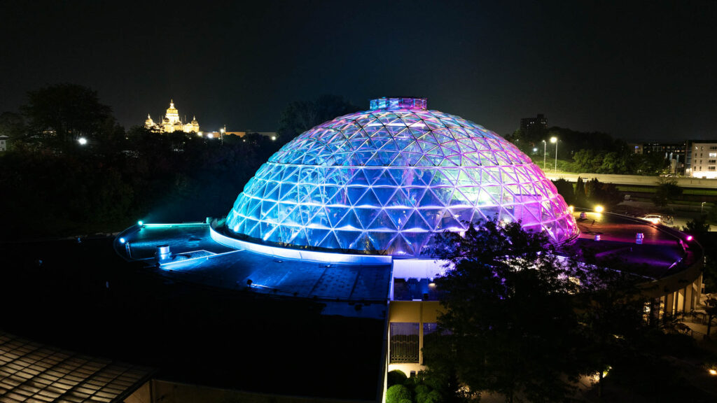 Des Moines Botanical Garden lit at night with multi-color cool tones and the Des Moines skyline in the background.
