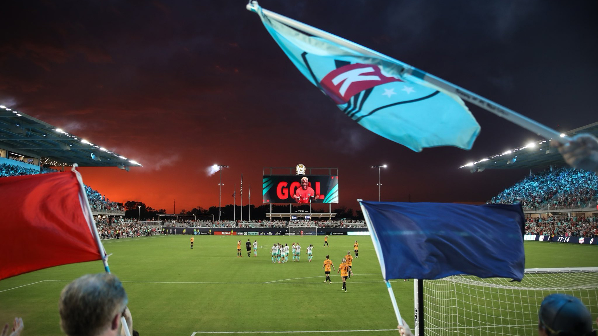 KC Current fans waving flags in the foreground with players on the LED lit soccer field.