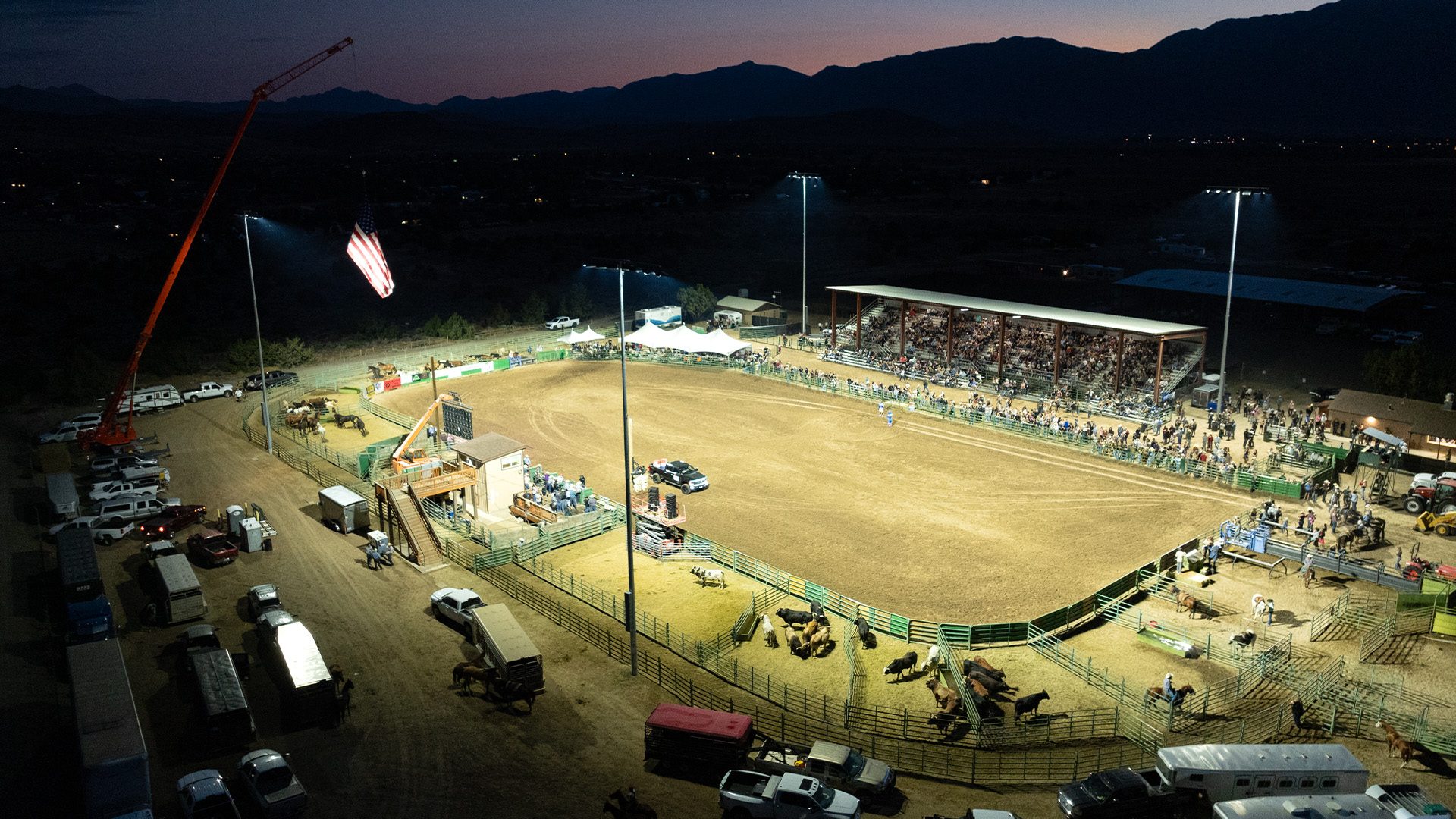 Rodeo arena lights turn on at Douglas County Fairgrounds as the sunsets. An American flag is flying from a crane on the left while spectators gather in the stands.